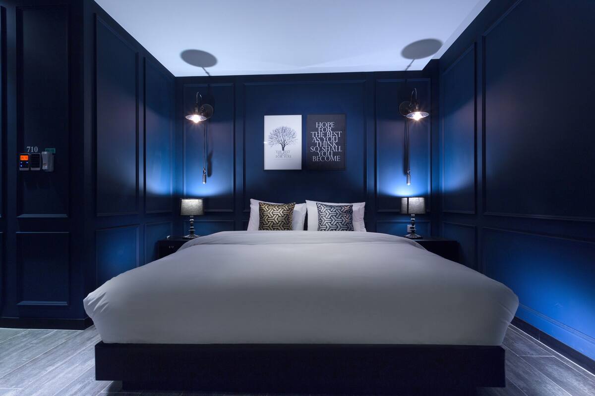 A large bed with a white mattress and two side tables, each with a lamp. There are two wall-mounted light fixtures and two framed inspirational quotes on the wall.
