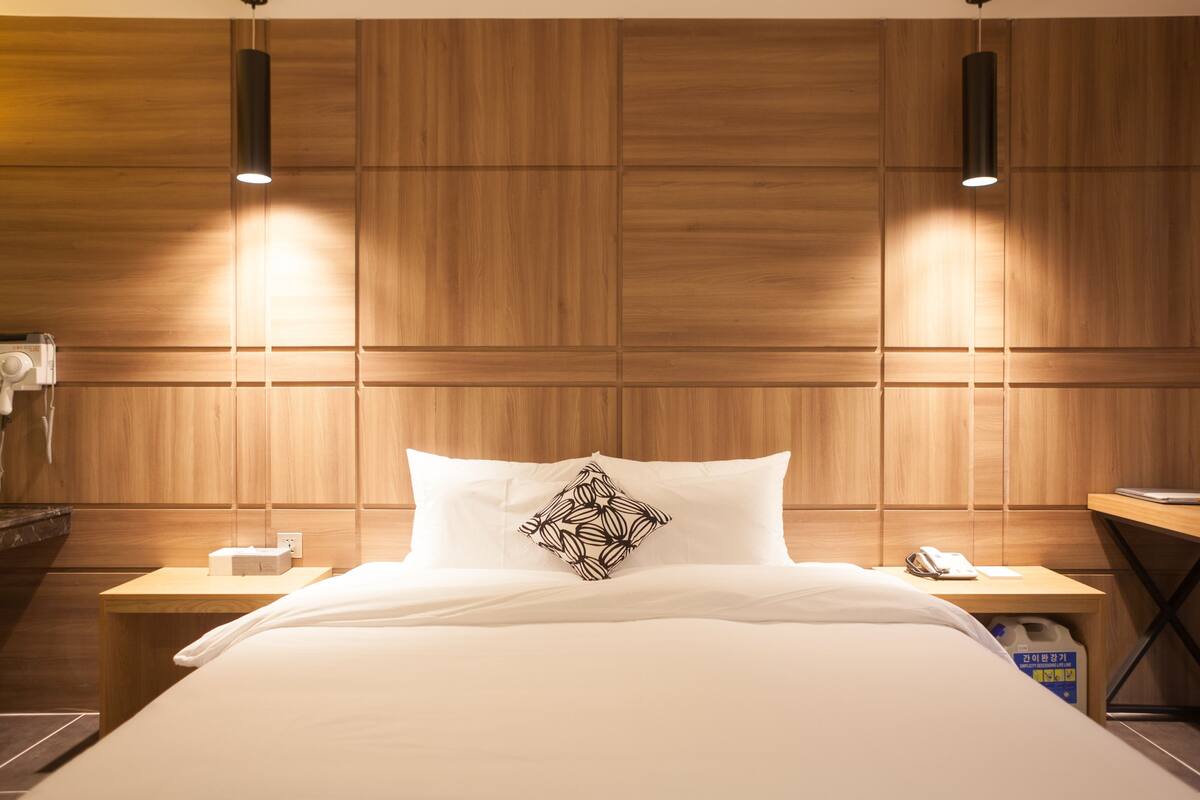 A hotel room with a wooden headboard, a bed with white bedding, a decorative pillow, bedside tables with lamps, and a nightstand with a phone and a tissue box.