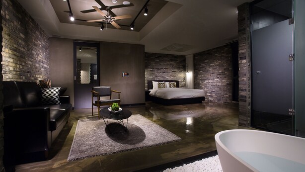 A modern bedroom with a large bed, a sitting area with a sofa and armchair, a small table with a plant, a ceiling fan, and a glass-enclosed shower area. - Hotel Sienna (Paju)