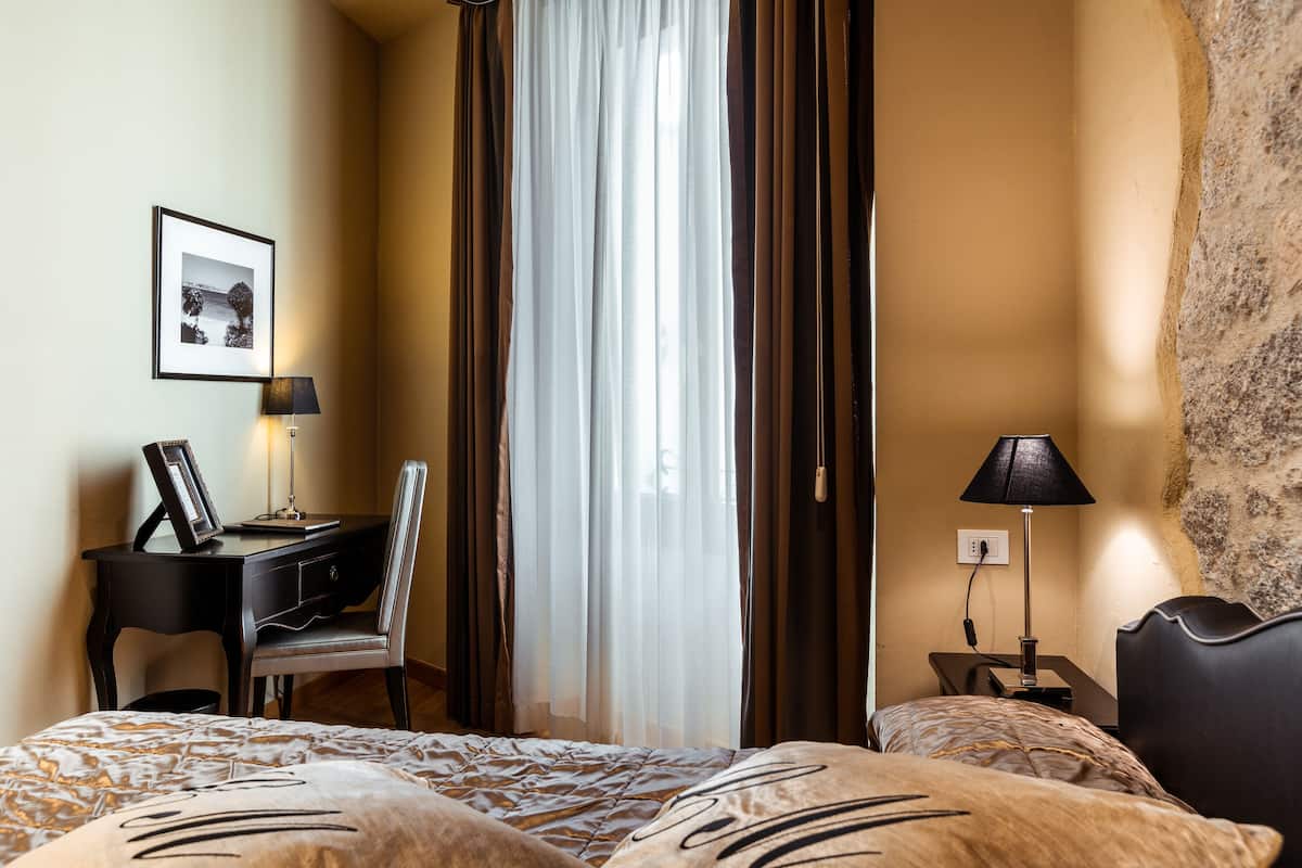A hotel room with a bed, a desk, a chair, a nightstand, a lamp, a picture on the wall, and a window with curtains.
