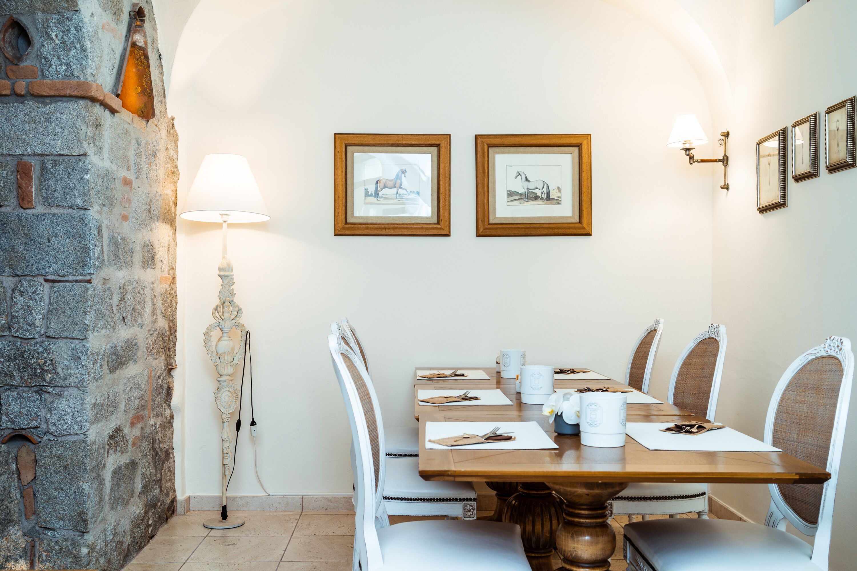 Un comedor con pared de piedra, una mesa con sillas blancas, cuadros en la pared y una lámpara de pie.