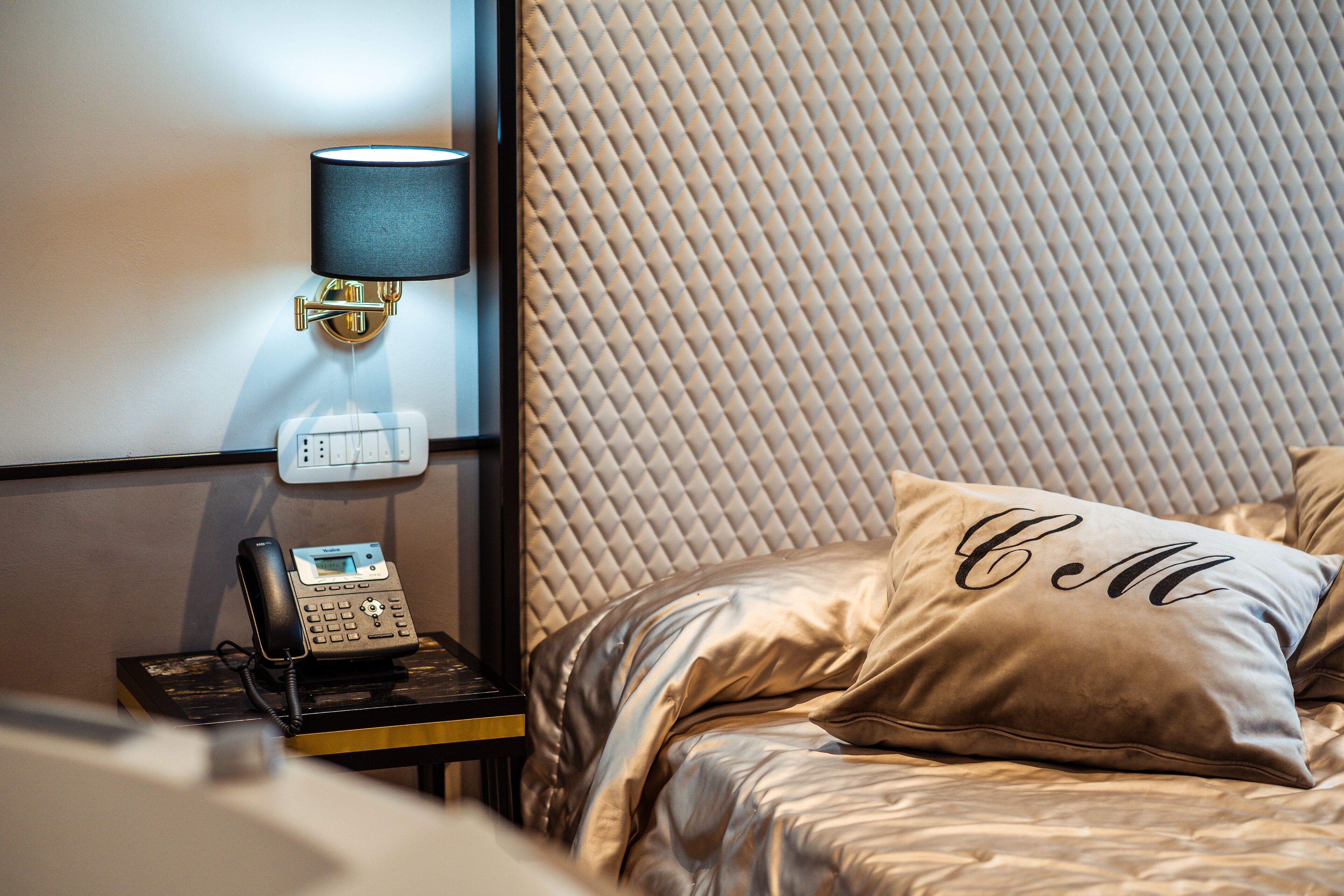 Habitación de hotel con cama, mesita de noche con lámpara, teléfono y cabecero texturizado.