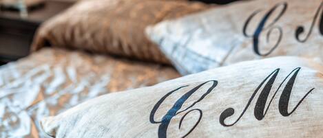 A bed with a headboard, pillows with embroidered initials, and a lamp on a bedside table.