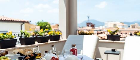 A breakfast spread on a terrace with a city view, including various cheeses, meats, and bread.