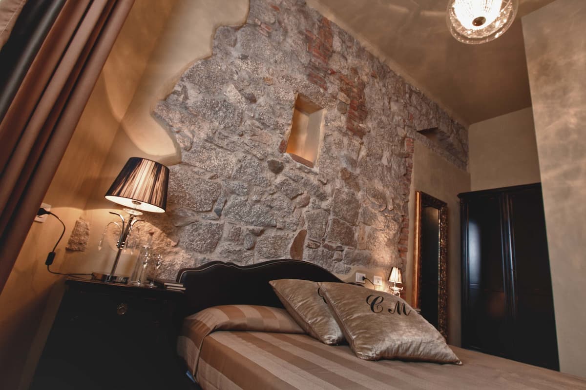 A bedroom with a stone wall, a bed with striped bedding, a nightstand with a lamp, and a door.