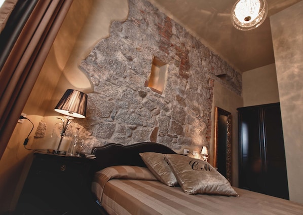 A bedroom with a stone wall, a bed with striped bedding, a nightstand with a lamp, and a door. - La Locanda del Conte Mameli (Olbia)