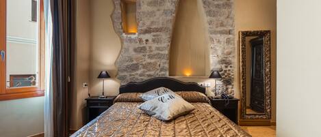 A bedroom with a stone wall, a bed with a patterned bedspread, a nightstand with a lamp, and a mirror with a decorative frame.