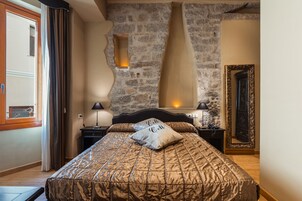 Un dormitorio con pared de piedra, una cama con colcha estampada, una mesita de noche con lámpara y un espejo con marco decorativo.