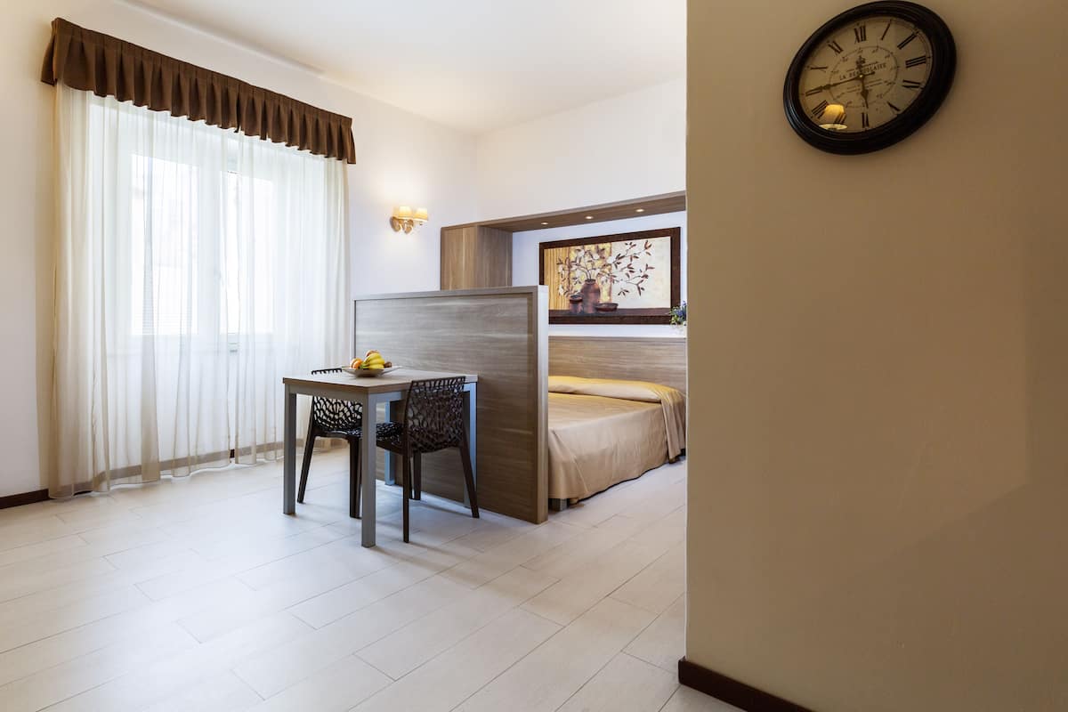 A hotel room with a bed, a dining table with chairs, a clock on the wall, and a window with curtains.