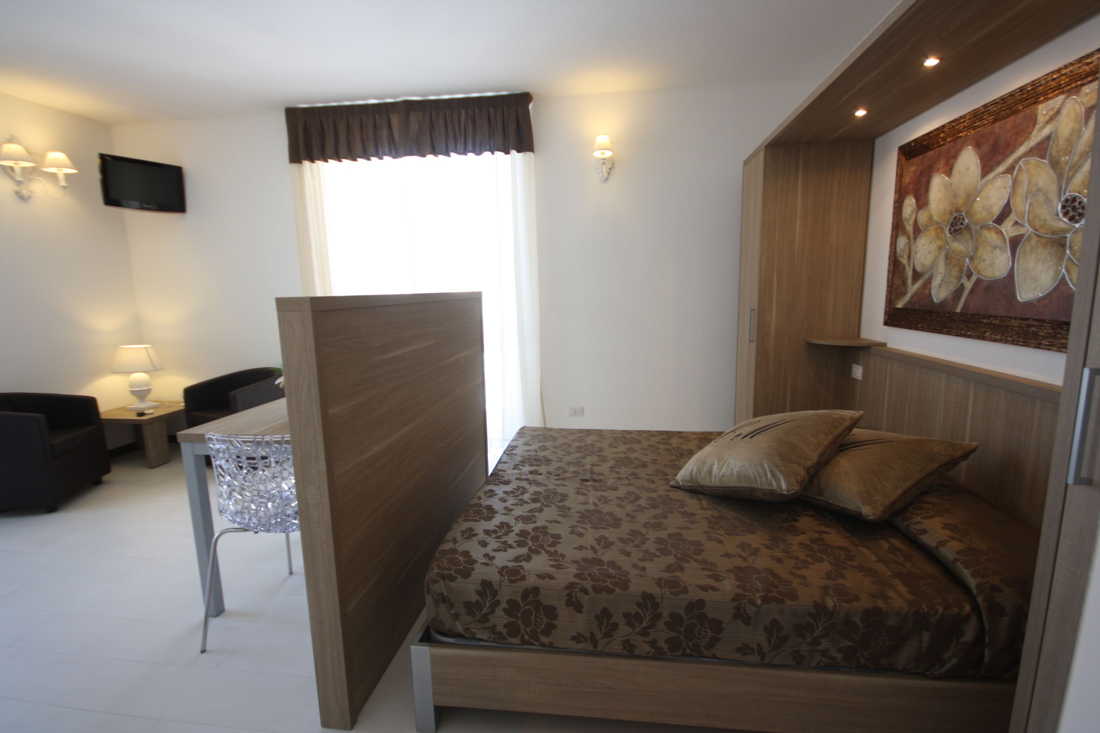 A bedroom with a bed, a TV mounted on the wall, a window with curtains, a small table with a lamp, and a painting on the wall.