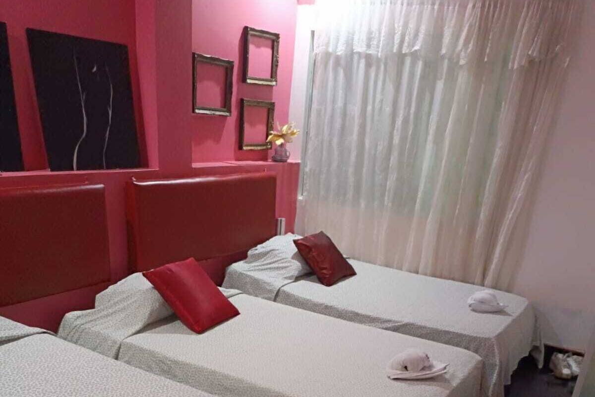 A hotel room with two beds, each with a white bedspread and a red pillow. There are framed pictures on the wall and a white curtain covering the window.