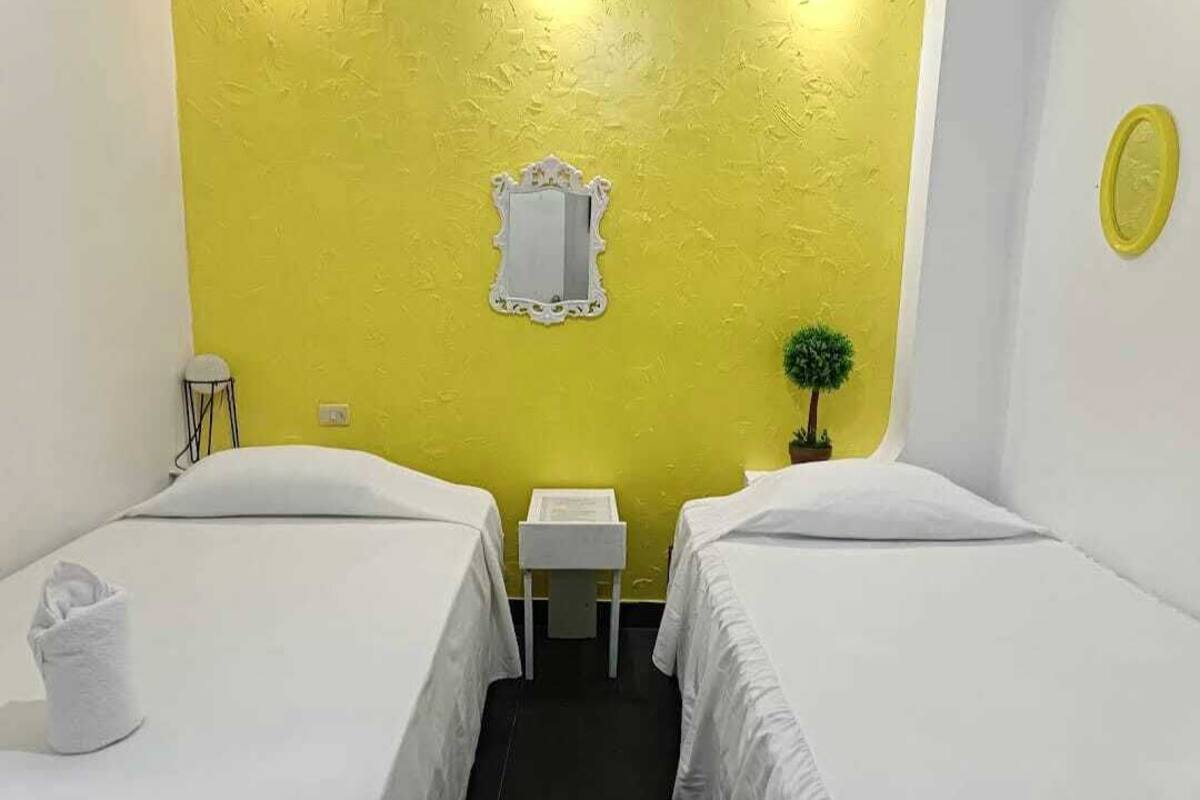 A hotel room with two beds, a small bedside table, a mirror, a potted plant, and a wall-mounted light fixture.