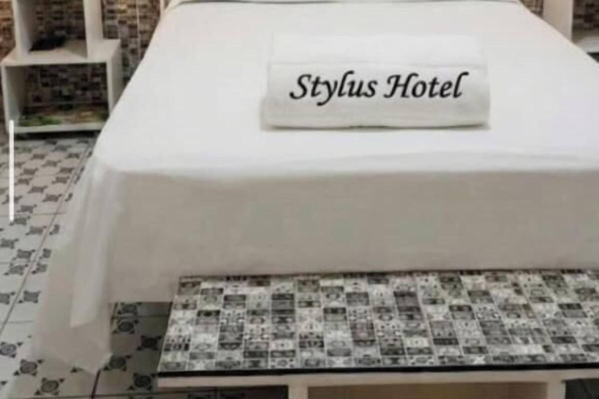 A hotel room with a bed, two bedside tables with lamps, a wall with Eiffel Tower silhouette, and a pillow with 'Stylus Hotel' text.
