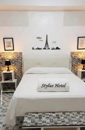 Une chambre d’hôtel avec un lit, deux tables de chevet avec des lampes, un mur orné de la silhouette de la Tour Eiffel et un oreiller avec l’inscription « Stylus Hotel ».