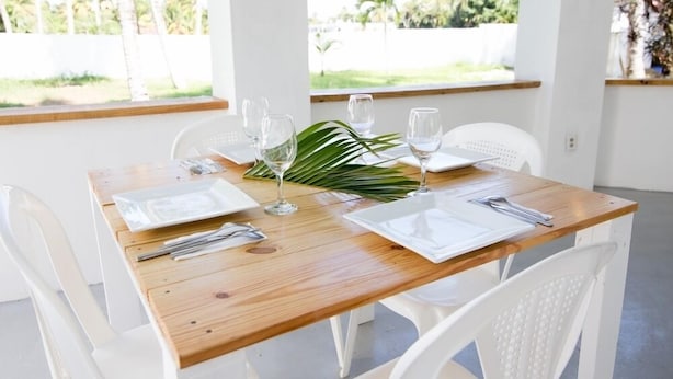 A dining table set with white plates, cutlery, and wine glasses, surrounded by white chairs, with a view of palm trees through large windows. - Northway's Hotel Monte Cristo (Cabarete)