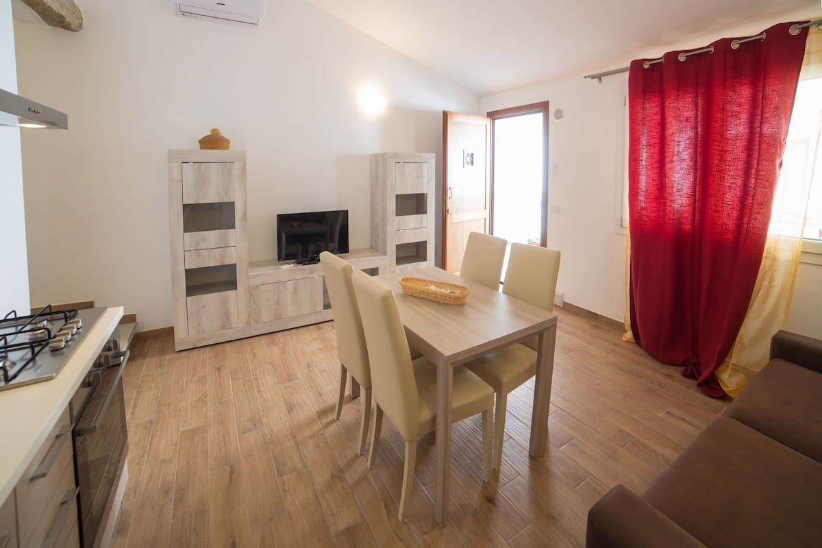 A modern kitchen with a gas stove, wooden cabinets, and a TV. There is a dining area with a table and chairs, and a red curtain on the window.