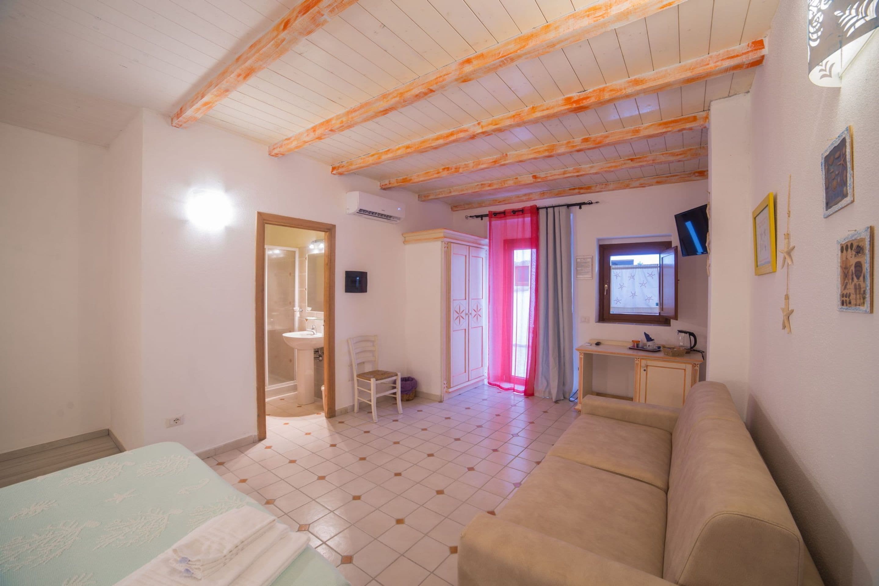 A room with a bed, a sofa, a small table, a chair, a television, and a bathroom visible through an open door.