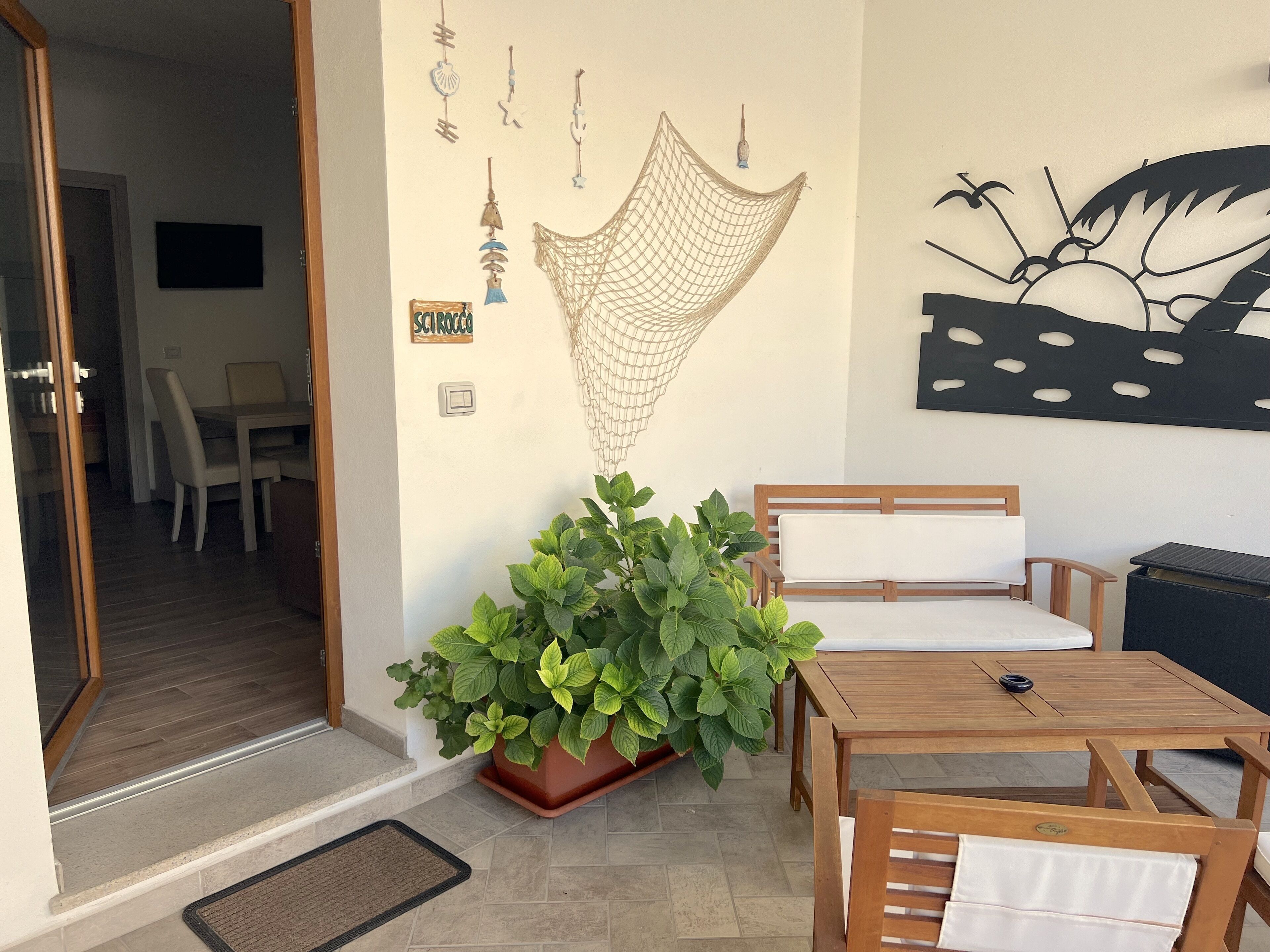 Un espacio de sala acogedor con una mesa y sillas de madera, una planta grande en maceta y adornos en la pared, incluyendo una hamaca y una obra de arte.