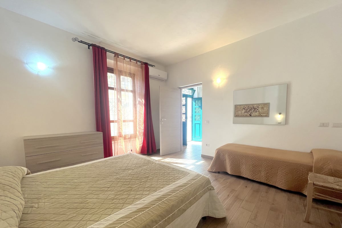 A bedroom with a bed, a dresser, and a chair. There is a window with curtains and a door leading to another room.
