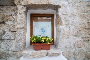 Un mur de pierre avec une fenêtre encadrée de bois, un rideau blanc et une jardinière remplie de fleurs jaunes.