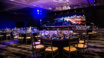 A banquet hall with round tables set for a formal event, a stage with a cityscape backdrop, and a bar area.