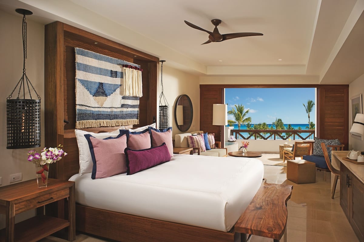A spacious bedroom with a large bed, a wooden headboard, a ceiling fan, and a balcony with a view of the ocean.