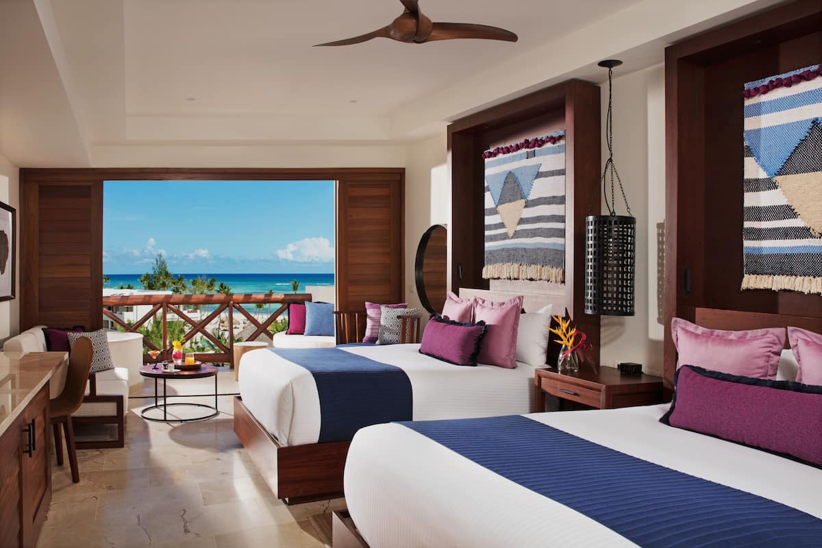 A hotel room with two beds, a balcony with a view of the ocean, a ceiling fan, and a wooden floor.