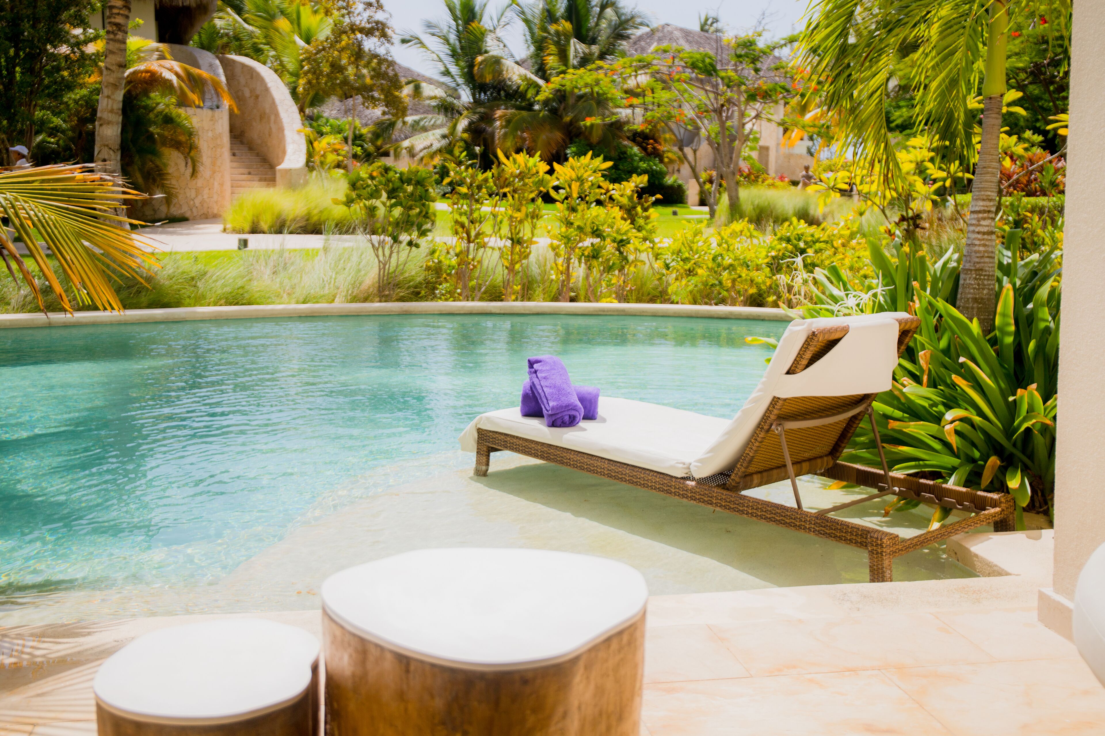 A pool area with a lounge chair and ottoman, surrounded by lush greenery and a stone structure.