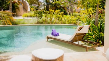A pool area with a lounge chair and ottoman, surrounded by lush greenery and a stone structure.