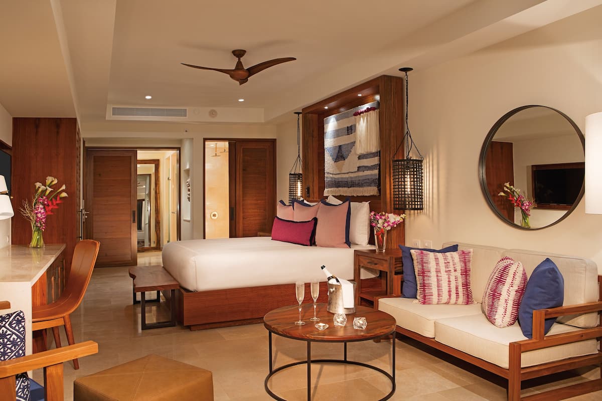 A hotel room with a bed, a sofa, a dining table, and a coffee table. There is a ceiling fan, a mirror, and a TV.