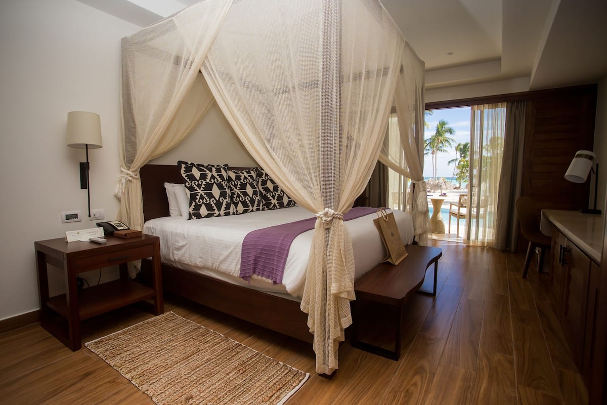 A bedroom with a bed, bedside table, lamp, and a view of the beach.
