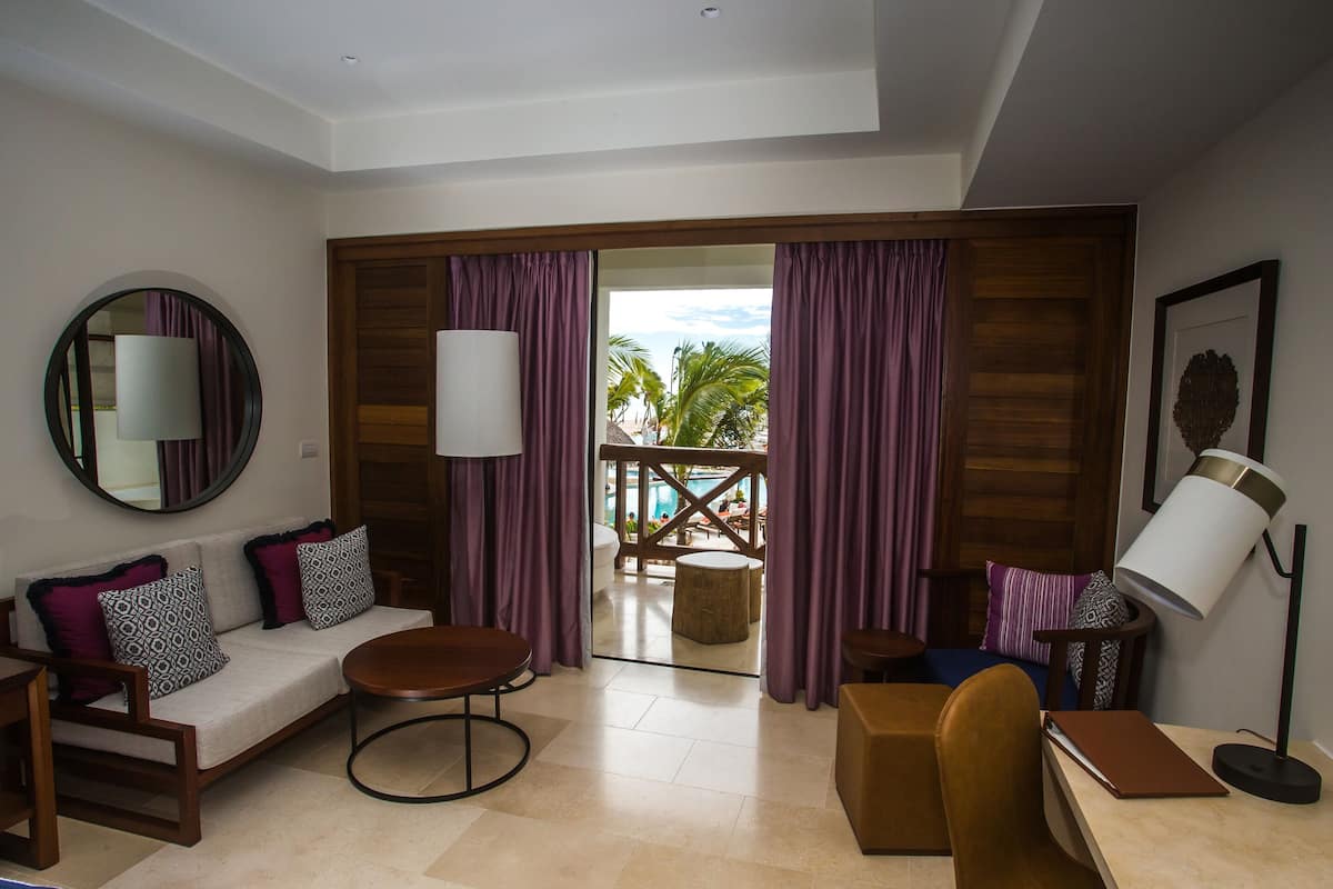 A modern hotel room with a sofa, round coffee table, and a desk with a lamp. A large window with purple curtains offers a view of the outdoors.