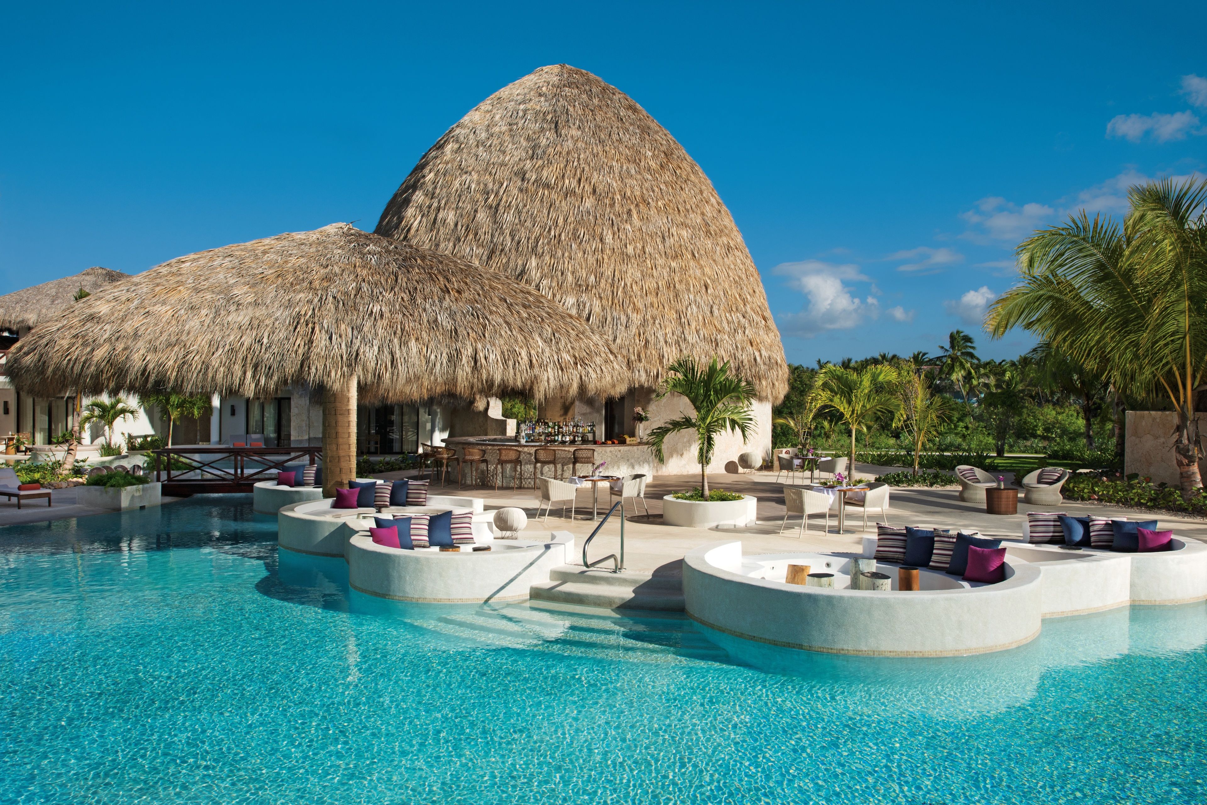 A resort with a large pool, outdoor seating areas, and a thatched-roof building.