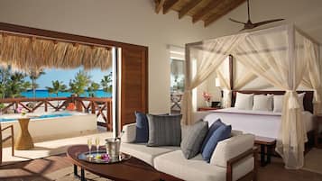 A hotel room with a bed, a sofa, a dining table with a tray of food, a small table with drinks, and a view of the ocean and palm trees.