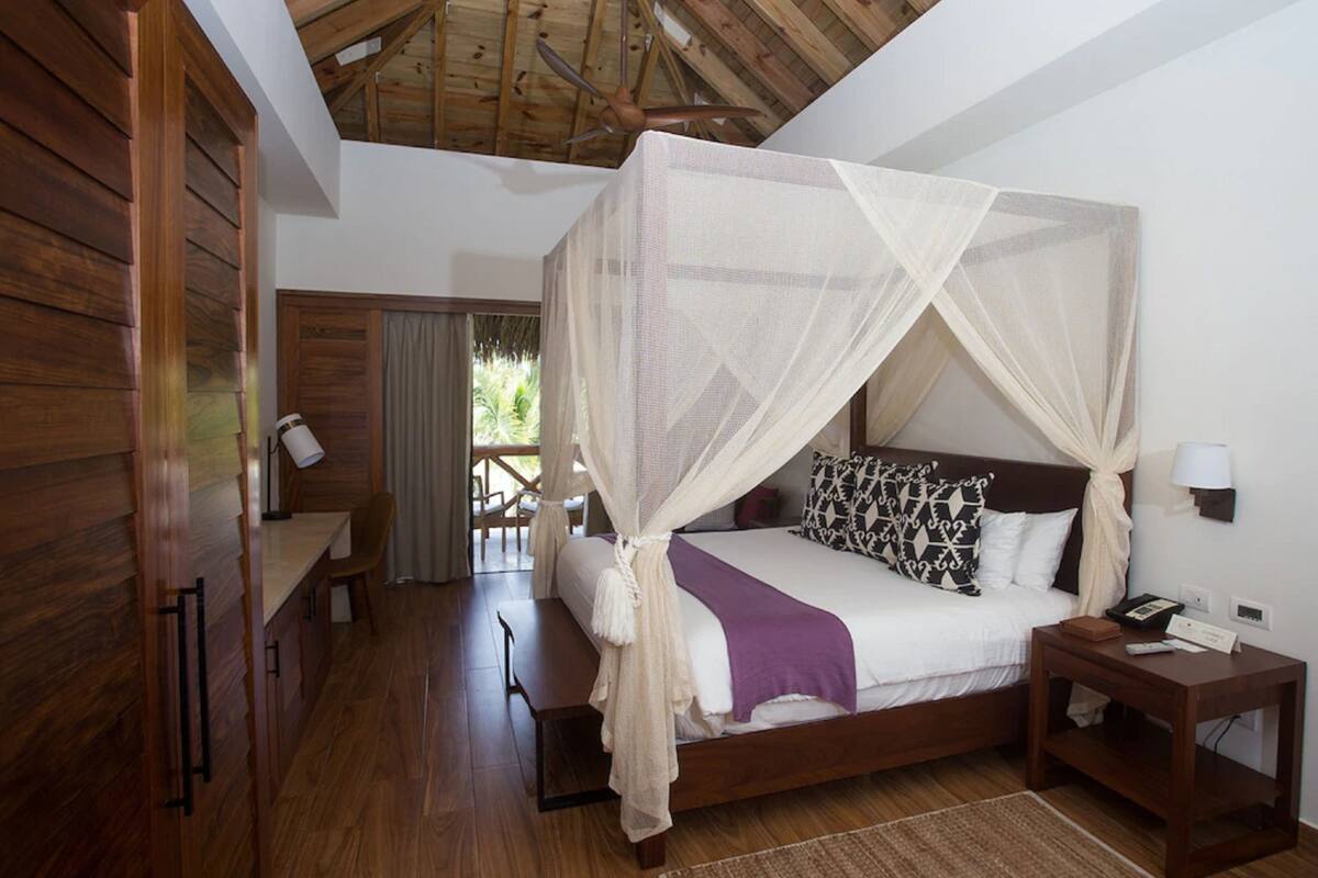 A bedroom with a wooden ceiling, a bed with a canopy, a bedside table, and a chair.