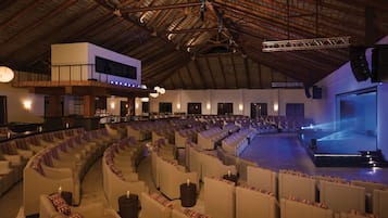 Un auditorium intérieur doté de gradins, d’une scène et d’un plafond recouvert de chaume.