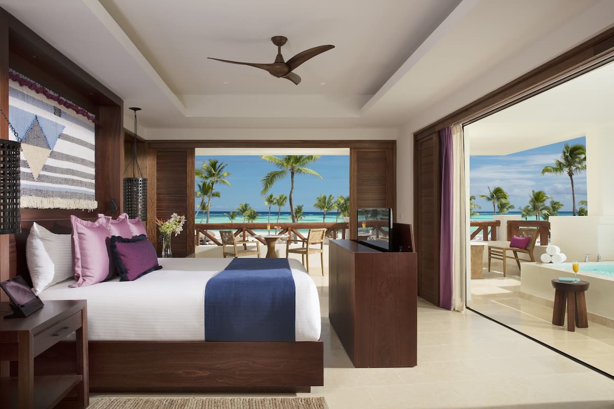 A bedroom with a large bed, a ceiling fan, a view of the ocean, and a balcony with a pool.