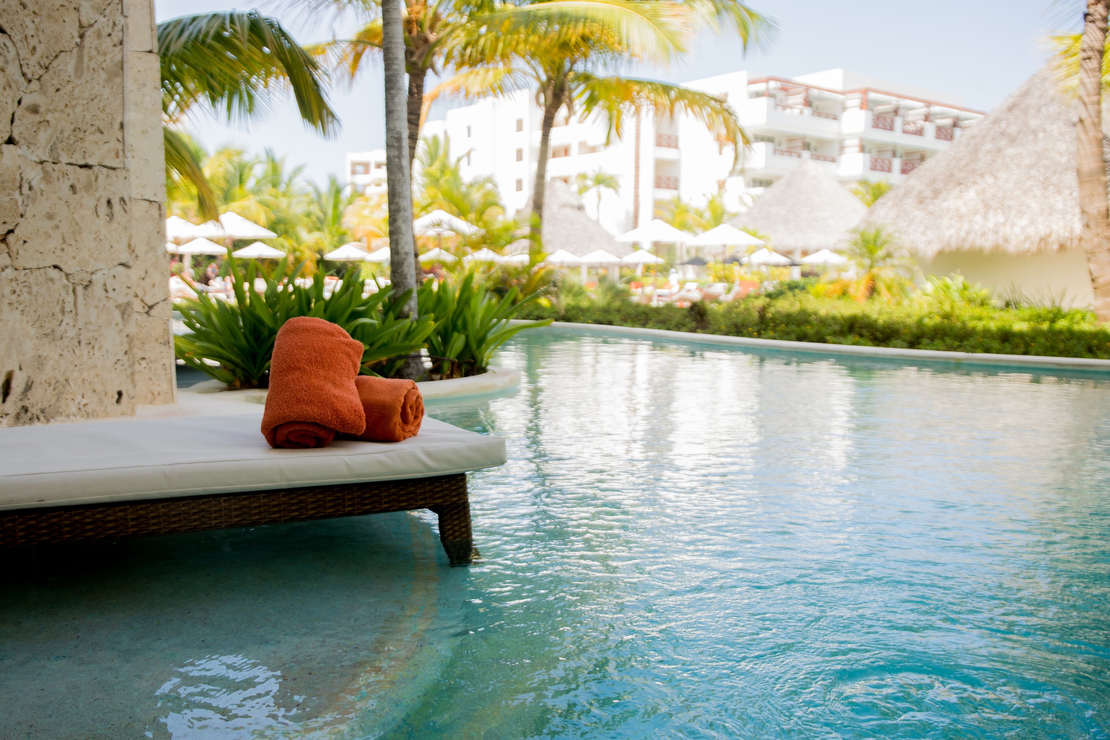 A poolside area with a lounge chair and a towel, surrounded by palm trees and a building with balconies.