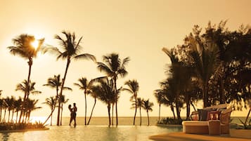 A couple standing by a pool with palm trees and a beach view.