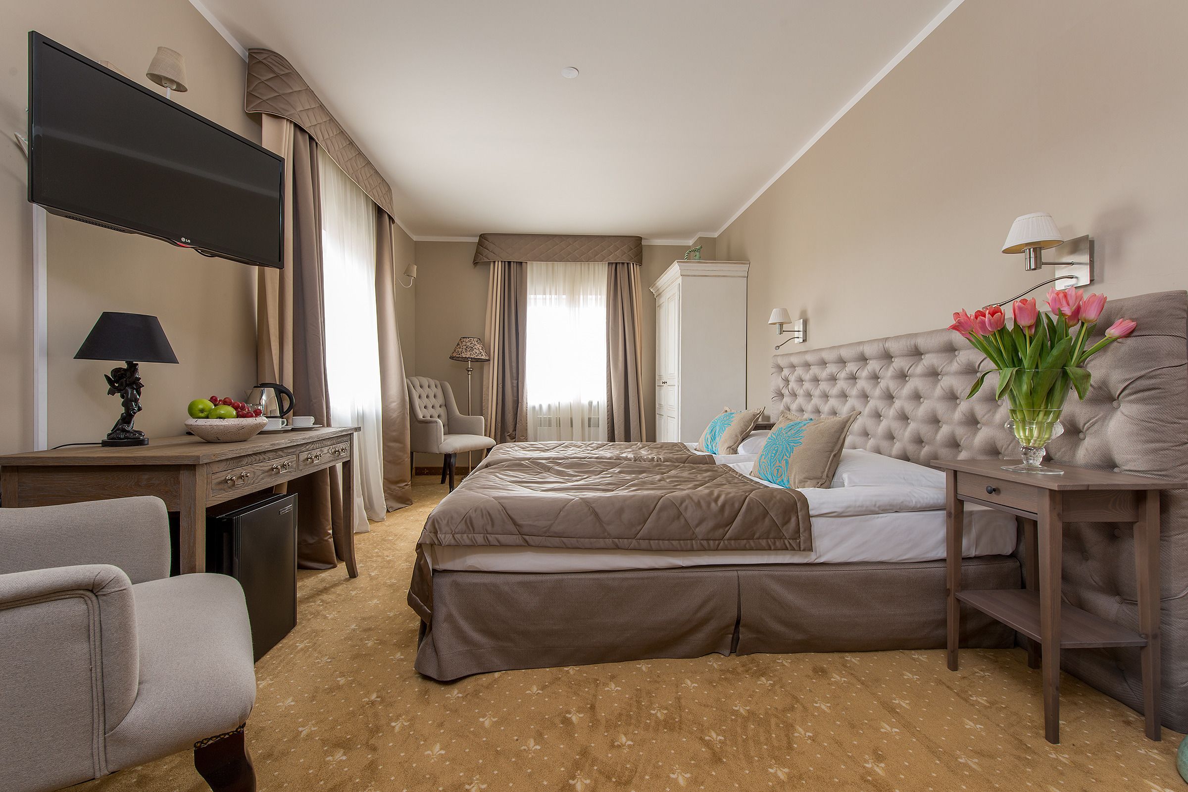 A hotel room with a bed, a desk with a lamp, a chair, a TV, and a vase with flowers.