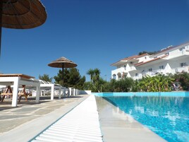 Un área de piscina con una piscina azul cristalina, sillas de descanso blancas y sombrillas.