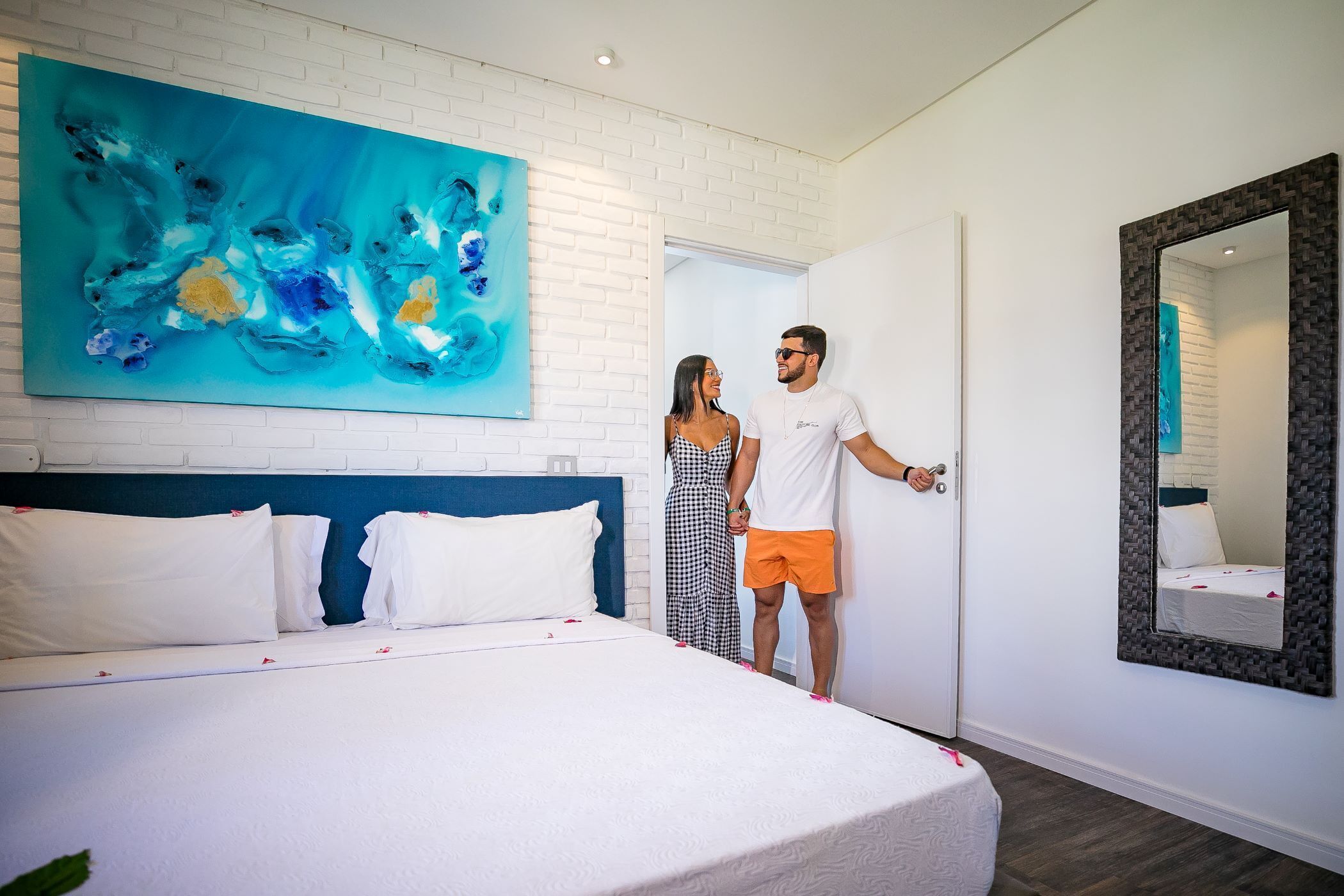 A hotel room with a large bed, a white brick wall, a blue abstract painting, and a mirror with a black frame.