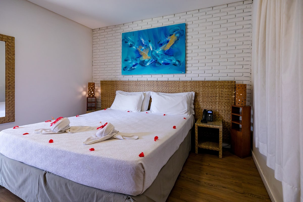 Een hotelkamer met een groot bed, witte linnens, een blauw abstract schilderij aan de muur en een houten hoofdeinde.