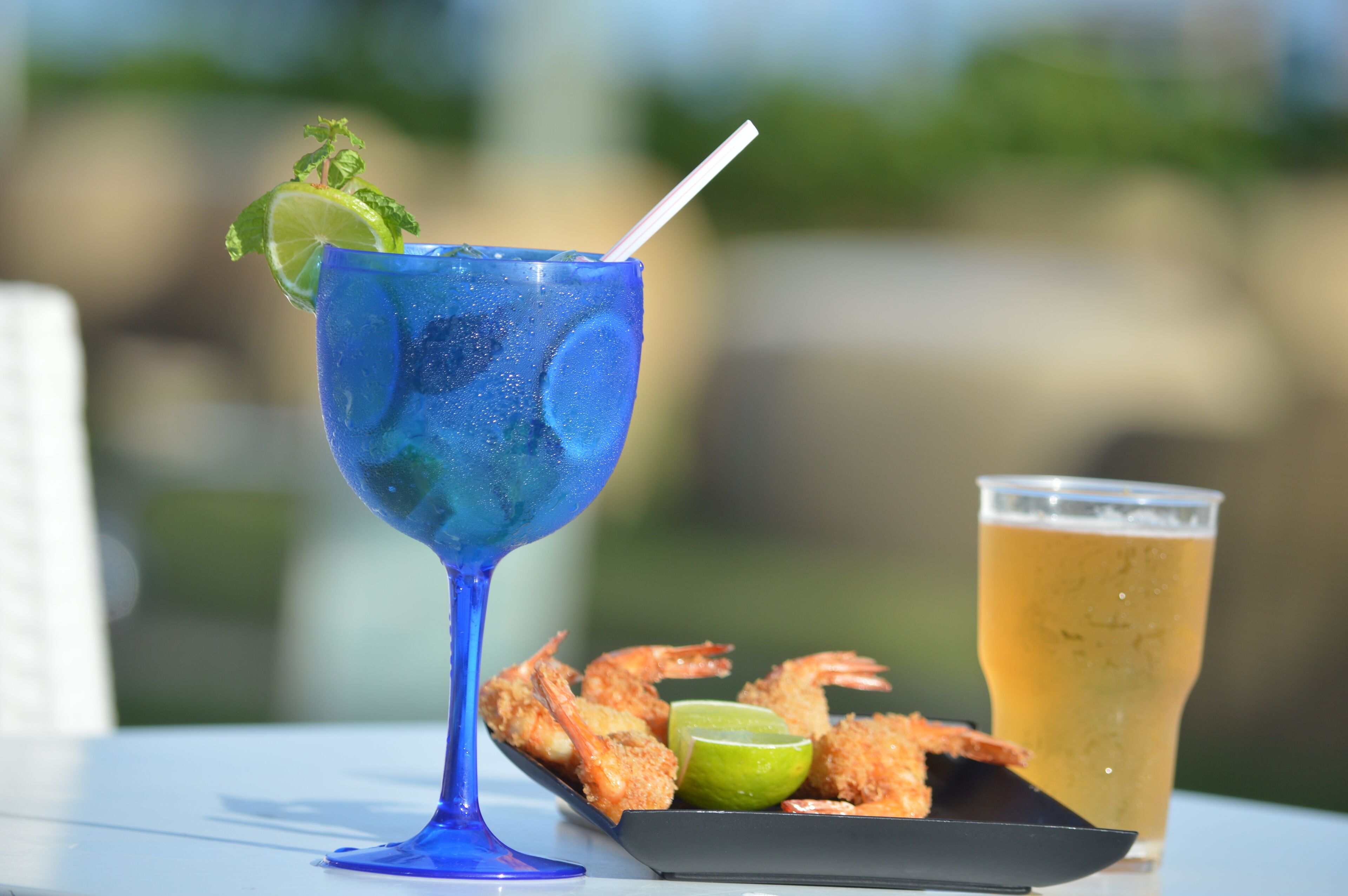 A blue cocktail glass with a lime wedge and mint, a plate of fried shrimp, and a glass of beer on a table.