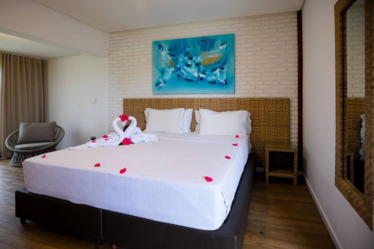 Een hotelkamer met een groot bed, een rieten stoel, een spiegel en een blauw abstract schilderij aan de muur.