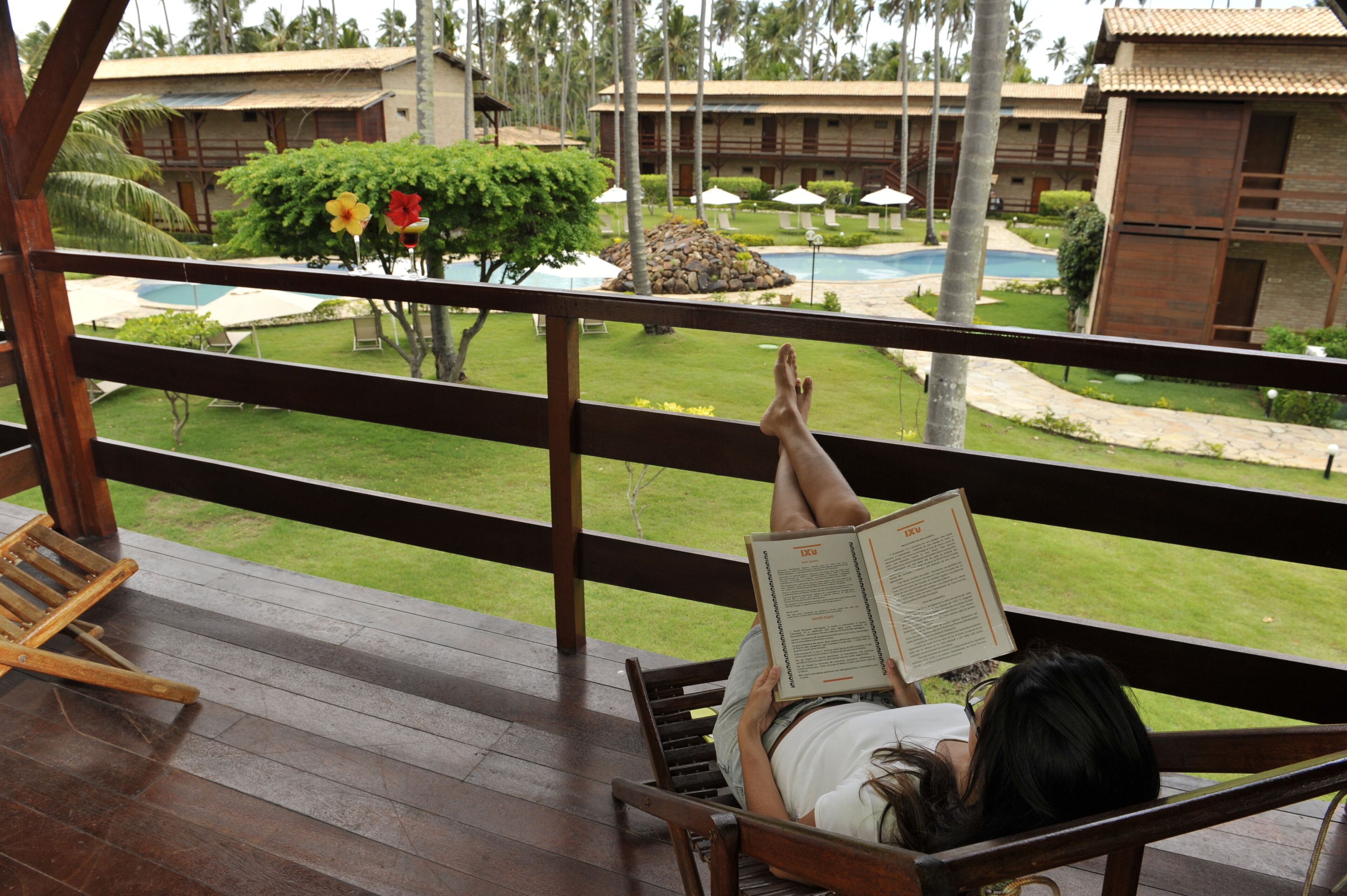 Una persona recostada en una silla de madera, leyendo un libro. La terraza da a una zona de piscina con sombrillas y un césped cuidado.