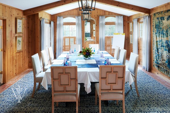 Uma sala de jantar com uma mesa comprida posta para oito pessoas, paredes de madeira e grandes janelas com cortinas.