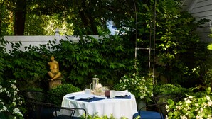 Ein runder Tisch mit weißem Tischtuch, umgeben von Metallstühlen, platziert in einem üppigen Garten mit dichter Bepflanzung und einer Statue im Hintergrund.