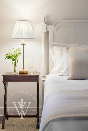 A neatly made bed with white linens, a bedside table with a lamp, a vase with flowers, and a clear glass bottle.