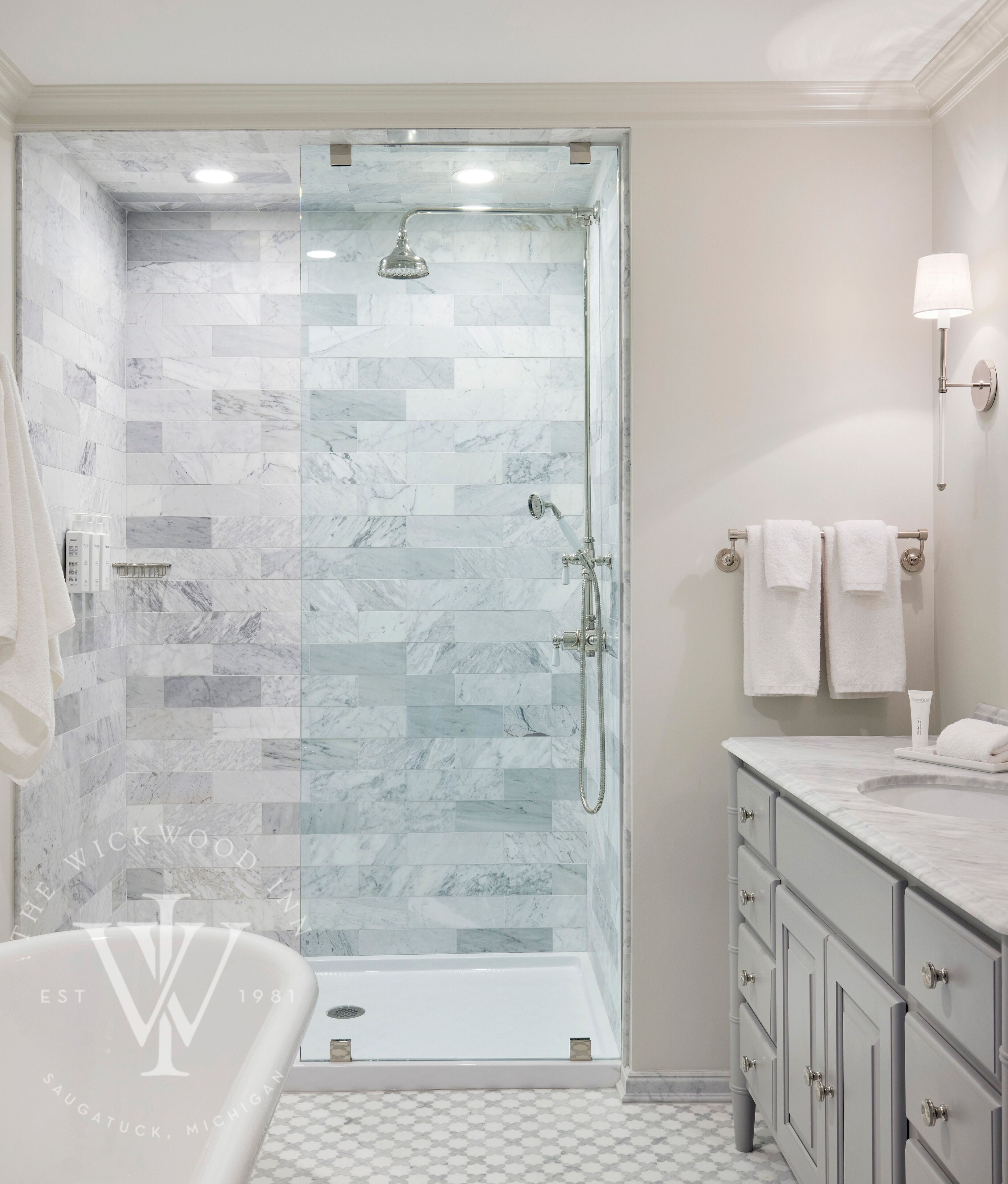 A modern bathroom with a glass-enclosed shower, marble tiles, a white bathtub, and a vanity with drawers.